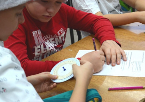Chłopiec trzyma koło szyfrujące i odkodowuje hasło. Drugi chłopiec zapisuje na kartce odszyfrowane litery.